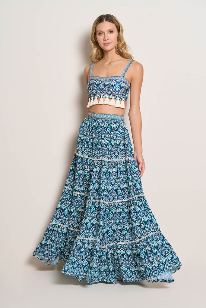 A model casually standing, wearing the Azure Bralette Bikini Top, with beautiful hand made tassels at the bottom of the lining, in white and blue colour combination, with adjustable straps, paired with a maxi skirt of the same pattern.
