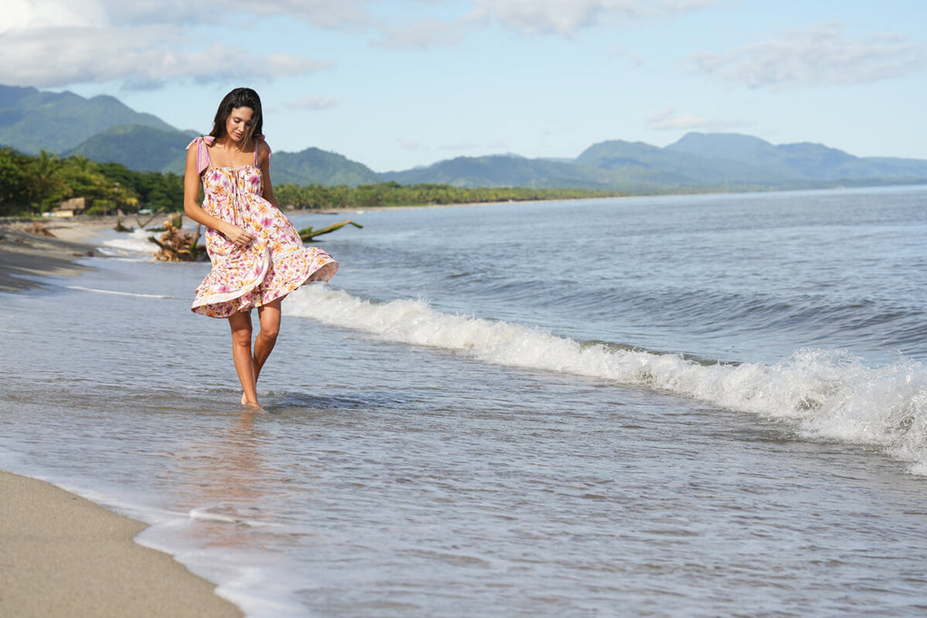 A model casually walking on the beachside, while wearing the Barbie Short Dress in white and pink flower print, with adjustable shoulder straps, a scrunch up on the rib section, handmade embroidery on the upper part of the double end ruffles.