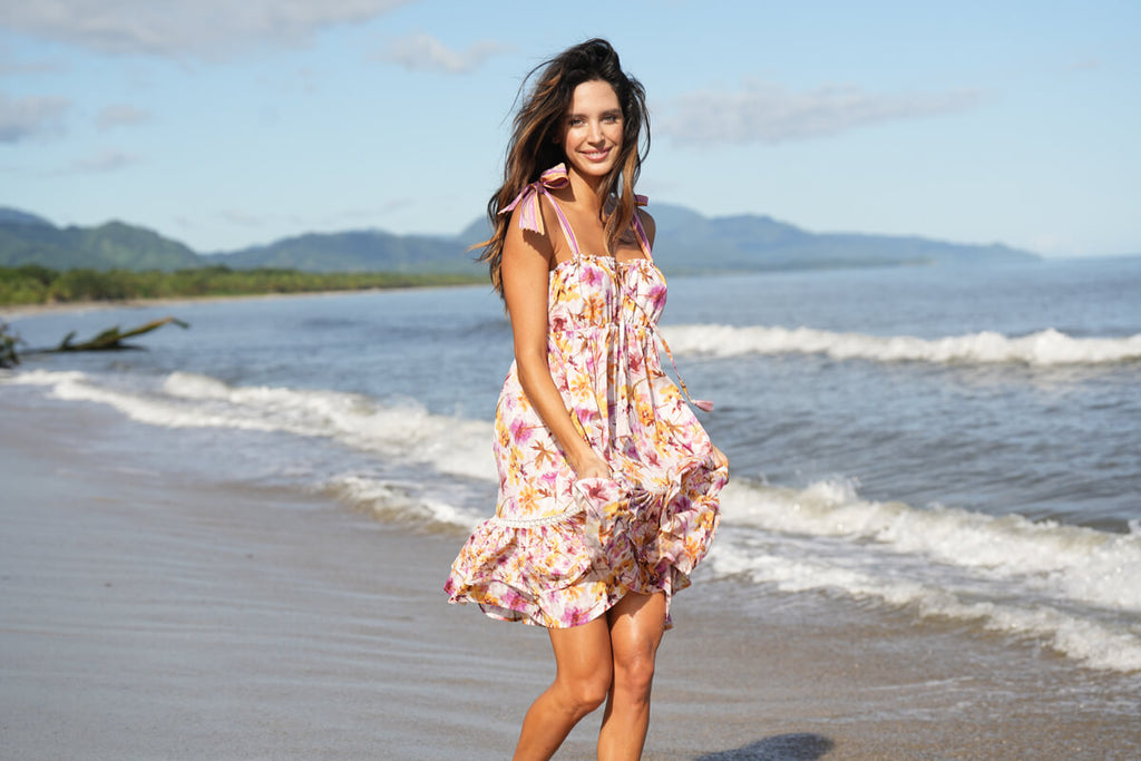 A model casually walking on the beachside, while wearing the Barbie Short Dress in white and pink flower print, with adjustable shoulder straps, a scrunch up on the rib section, handmade embroidery on the upper part of the double end ruffles.